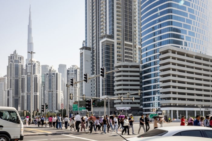 سقوط آرامش دبی؛ چگونه جنگ ایران تصویر لوکس خلیج فارس را بر هم زد؟ Pedestrians crossing a street in Dubai's Business Bay financial district, with the Burj Khalifa visible in the background.